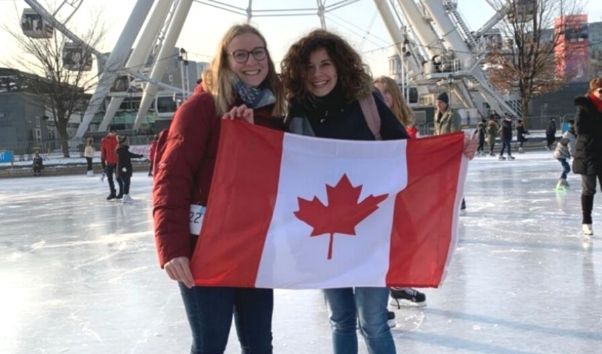 Austauschschülerinnen auf einer Eislaufbahn halten eine Kanada Flagge in die Luft. Im Hintergrund steht ein Riesenrad.